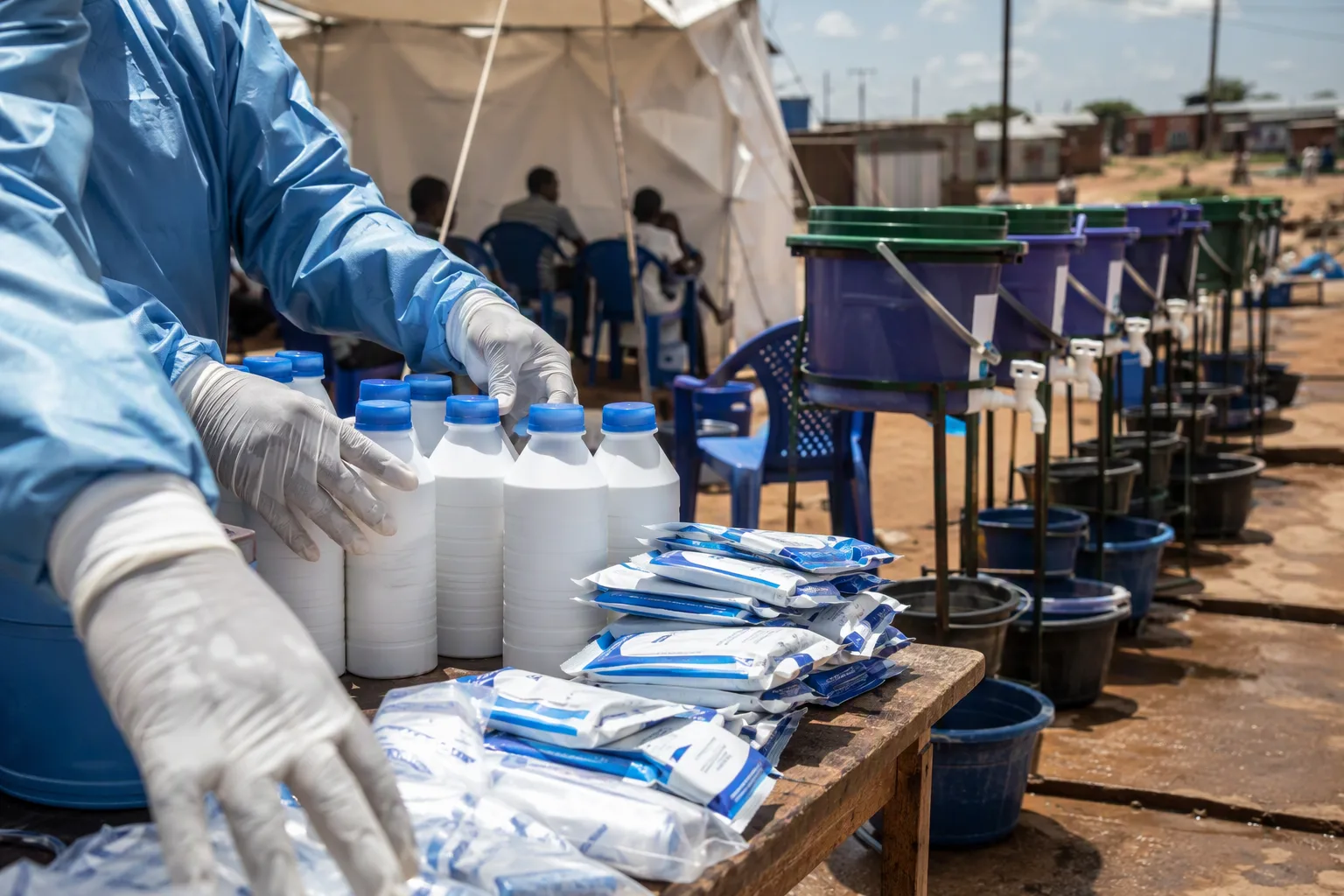 Gloved hands stacking chlorine bottles and oral rehydration sachets at a temporary cholera treatment tent